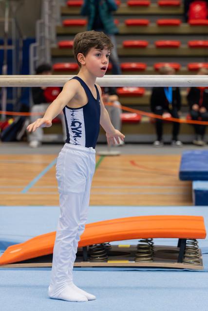 Young boy in navy leotard stands alert by springboard, arms slightly raised, focused expression in a gymnastics arena.