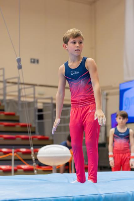 Young male gymnast in pink and navy leotard stands focused on blue mat, hands chalked, composing himself before routine.