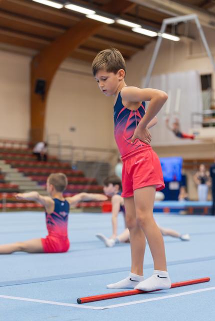Young boy in red and navy leotard stands focused on a floor rod, hands on hips, in a gymnastics hall.