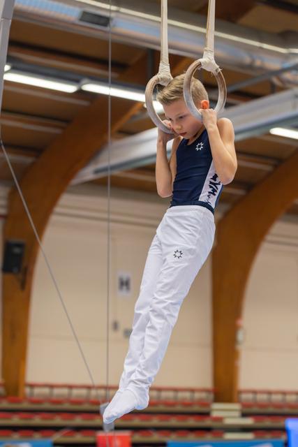 Focused young male gymnast grips still rings mid-routine, legs tucked, intense concentration in a gymnasium.