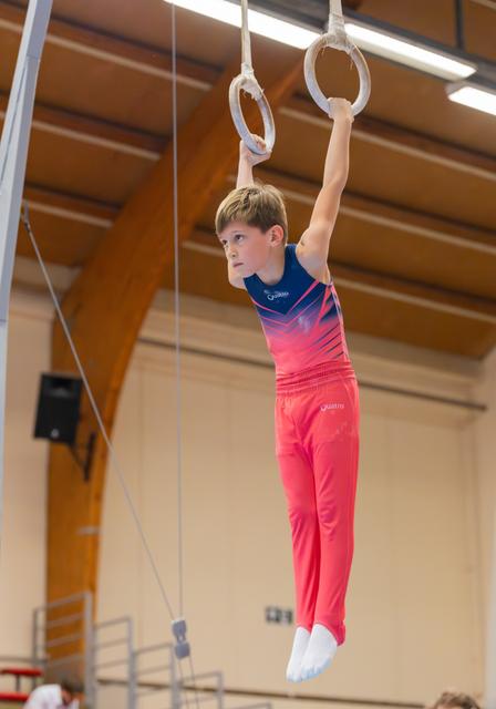 Determined young boy hangs from still rings in a gymnastics hall, wearing a red and blue Quattro leotard.