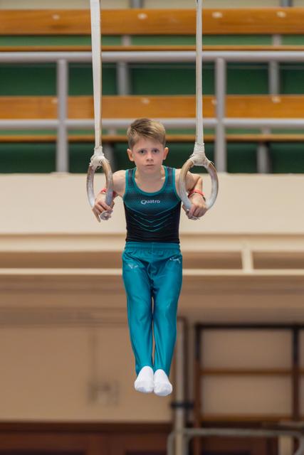 Focused young boy holds steady on gymnastics rings, wearing teal Quatro leotard, legs tucked, intense concentration on his face.