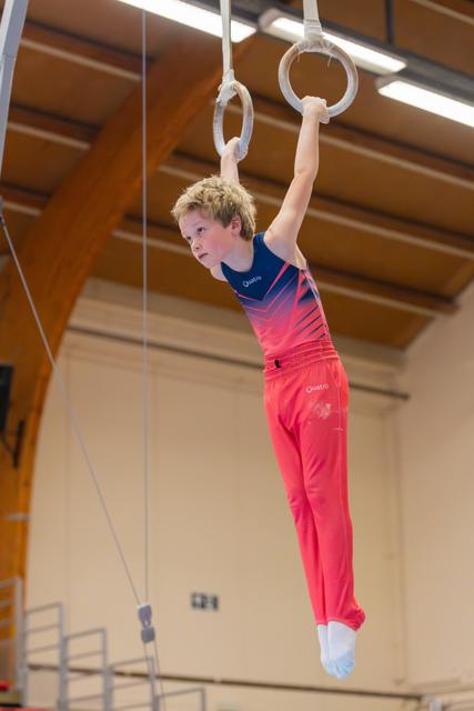 Young boy in red and blue Quattro kit hangs with intense focus on gymnastic rings in a sports hall.