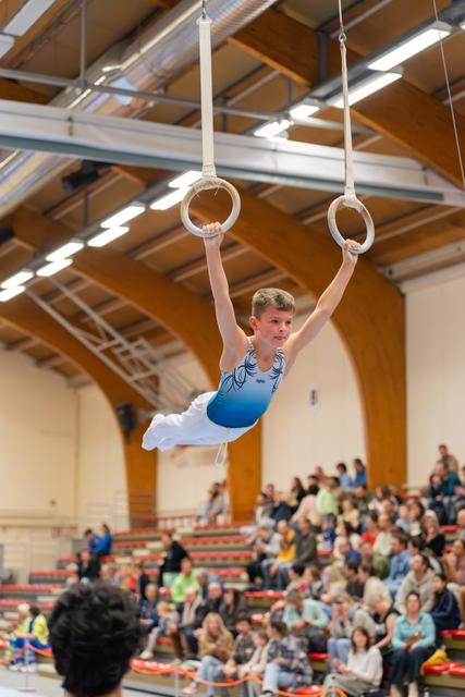 Young male gymnast hangs focused on still rings, body taut, in a packed indoor gymnasium with spectators watching.