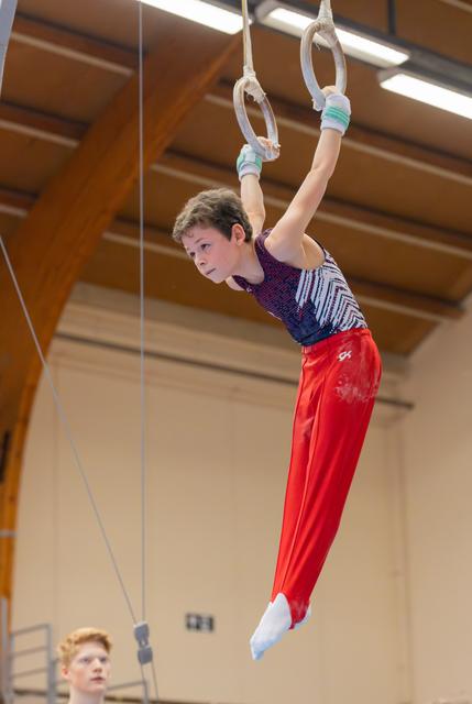 Young male gymnast hangs focused on still rings, red pants and wrist guards, in a wooden-ceiling gymnasium.