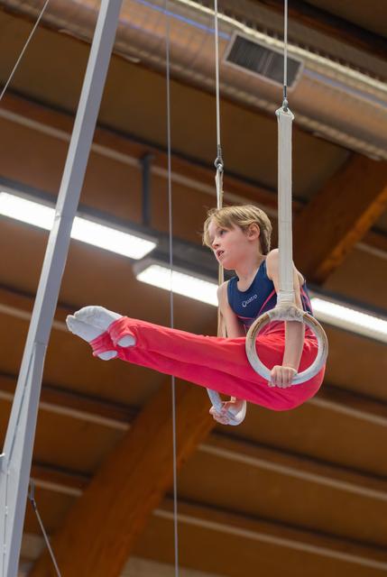 Focused young boy performs a straddle hold on gymnastics rings in an indoor sports hall, legs extended with intense concentration.