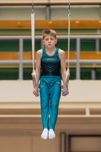 Young boy gymnast holds still rings with focused expression, wearing teal Quatro leotard in gymnasium