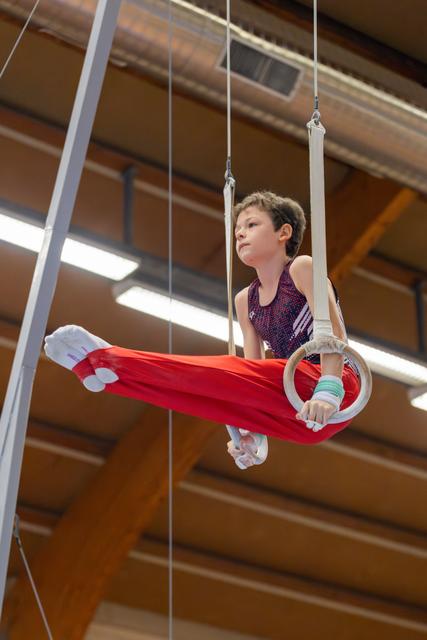 Determined young boy grips gymnastic rings mid-routine, legs extended, wearing red pants in an indoor gymnasium.