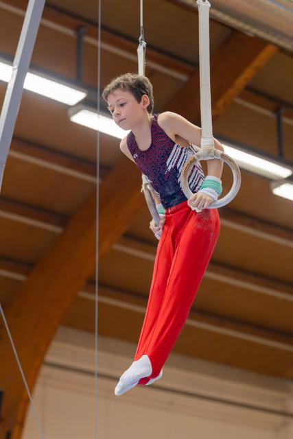 Focused young boy grips gymnastic rings mid-routine, body angled with concentration in a wooden-beamed sports hall.