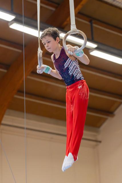 Young male gymnast hangs from still rings with intense focus, body angled, wearing red pants and sequined leotard indoors.