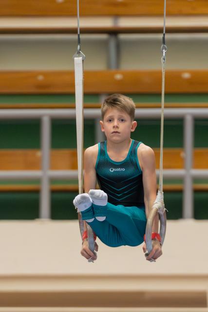 Young boy in teal Quatro leotard holds a tuck position on still rings, expression calm and concentrated in a gymnasium.