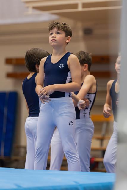 Young boy in navy leotard and white pants stands focused, gazing upward, awaiting his turn on the gymnastics floor.