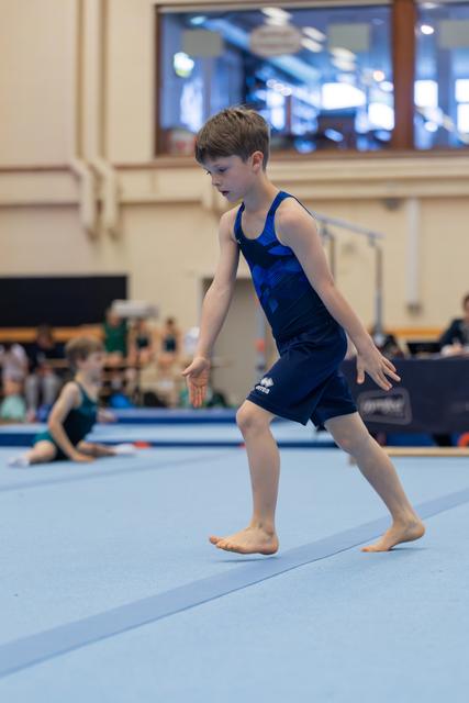 Young boy in blue leotard walks across the floor mat with focused concentration during a gymnastics event.
