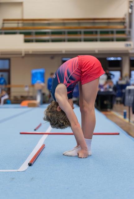 Young gymnast bending forward in a deep toe touch on the blue floor exercise mat during an indoor gymnastics meet.