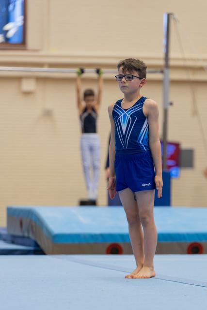 Boy in blue gymnastics leotard and glasses stands still, focused, on a blue mat while a teammate trains on bars behind.