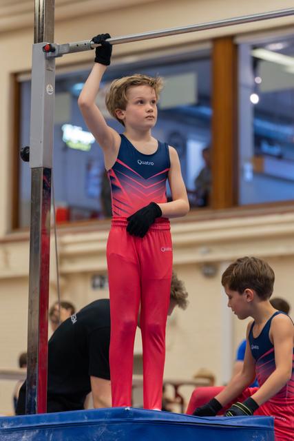 Focused boy in red Quatro leotard grips the high bar, standing tall, while a teammate waits nearby in a gym.