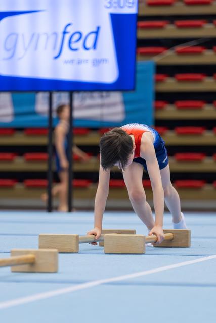 Young gymnast bent over wooden pommel handles on a blue mat, focused and preparing at a Gymfed event.