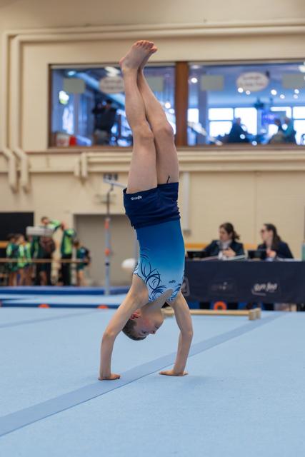 Young male gymnast performs a straight handstand on the floor exercise mat, legs perfectly vertical, wearing blue shorts.