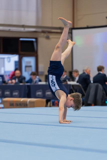 Young boy performs a precise handstand on the floor exercise mat, legs extended upward, during an indoor gymnastics competition.