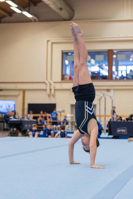 Shirtless boy performs a focused handstand on the blue floor mat during a gymnastics competition.