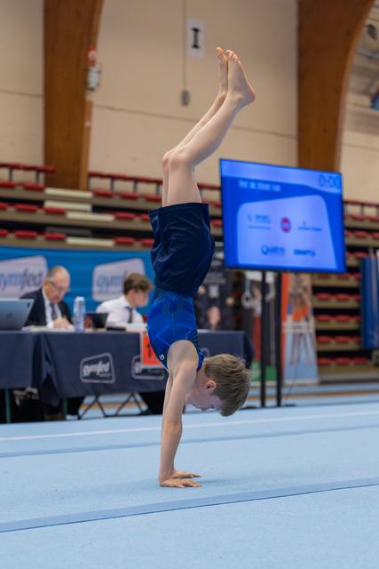 Young male gymnast performs a focused handstand on the floor mat during a gymnastics meet, judges visible behind.