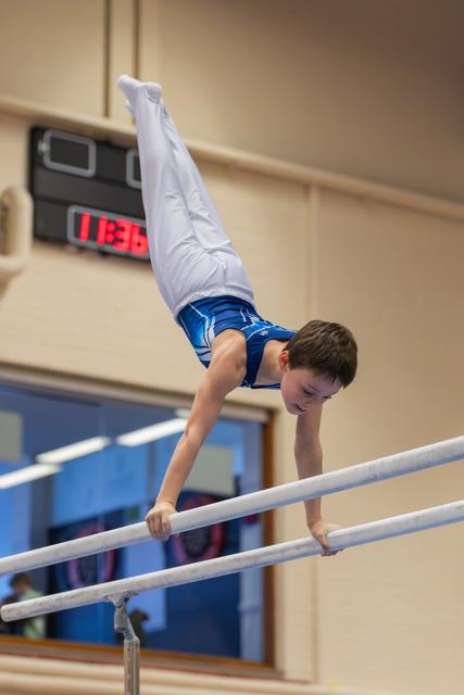 Young boy performs a handstand on parallel bars, legs split wide, face showing intense concentration in an indoor gym.