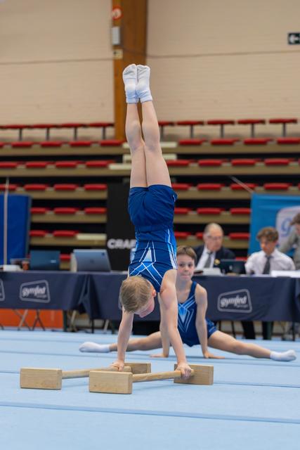 Young gymnast performs precise handstand on wooden blocks while teammate holds splits behind, judges watching in background.