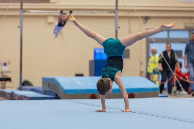 Young gymnast performs a perfect handstand with legs in a full split on a blue gymnastics mat during a competition.