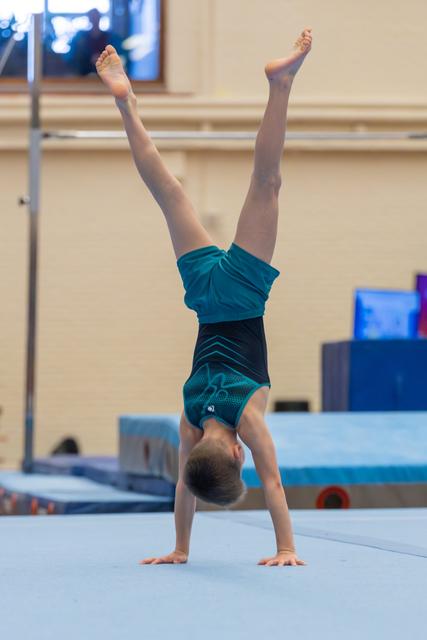 Young gymnast performs a perfect handstand on the floor exercise mat, legs split high in a teal competition leotard.