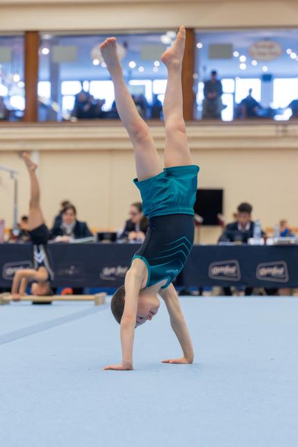 Young gymnast performs a focused handstand on the floor exercise mat, legs perfectly vertical, judges visible in background.