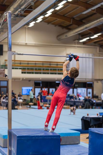 Young gymnast in red and navy leotard grips the high bar with both hands, standing on a blue block in an indoor gym.
