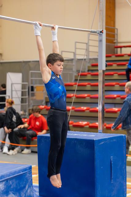 Focused young boy hangs from a horizontal bar in a blue leotard, feet lifted, competing in a gymnastics hall.