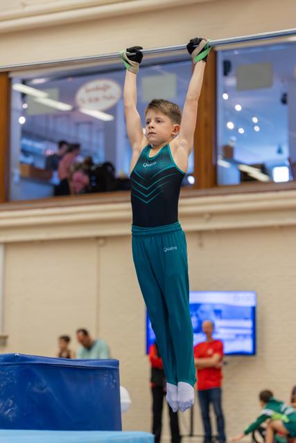 Young boy in teal Quatro leotard hangs from the high bar with focused concentration, arms fully extended.