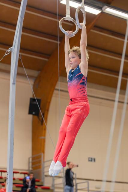 Young male gymnast hangs from still rings with focused expression, wearing red and blue leotard in a sports hall.