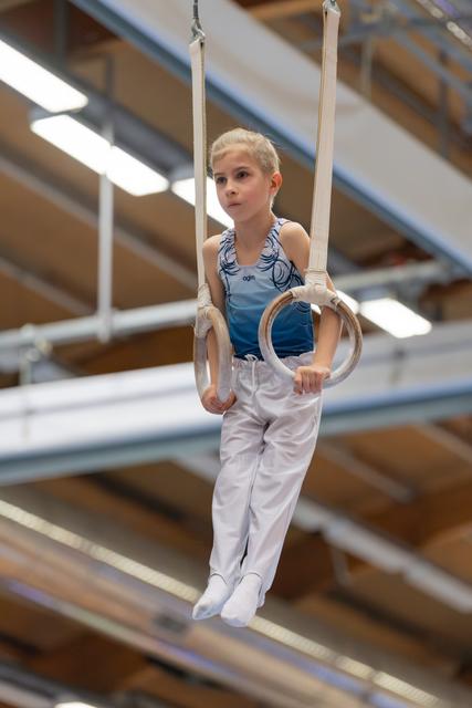 Young gymnast hangs calmly on still rings, expression focused and composed, inside a bright gymnastics hall.