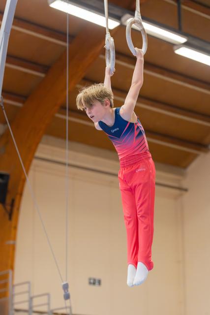 Young male gymnast hangs from still rings with focused expression, wearing red and blue Quatro leotard in a gymnasium.