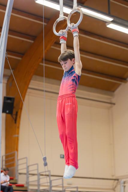 Focused young male gymnast hangs from still rings, looking down with concentration, in a wooden-beamed sports hall.