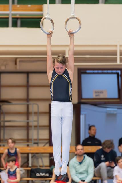 Young male gymnast hangs from still rings with focused concentration, arms extended overhead, in a gymnasium setting.