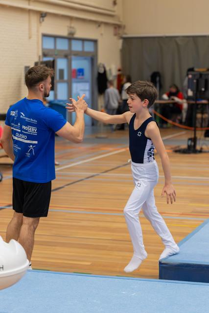 Young male gymnast leaps off vault to high-five his smiling coach in a bright gymnasium, radiating pride and encouragement.