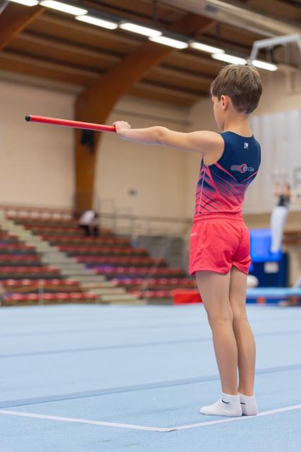 Young boy in red and navy leotard stands poised on blue mat, extending a red stick outward with focused posture.