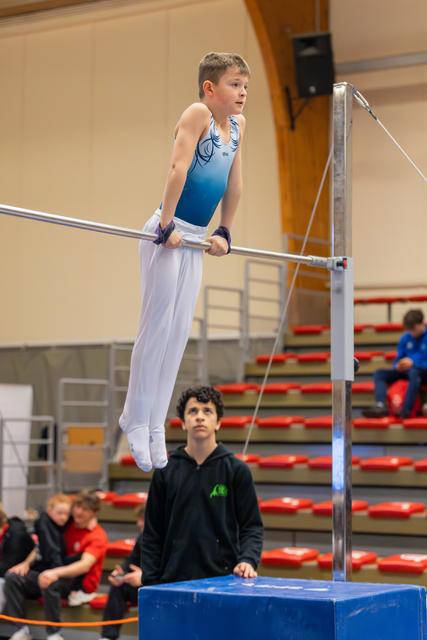 Young boy grips the high bar with focus, while a spotter in black hoodie watches closely from below in a gymnastics hall.