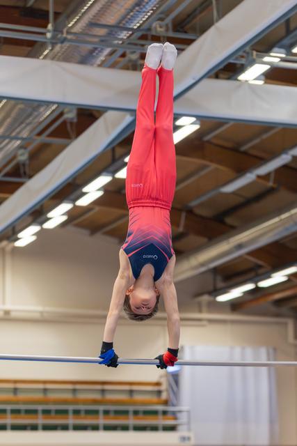 Young male gymnast performs an inverted handstand on the high bar, body fully extended, in an indoor gymnastics hall.