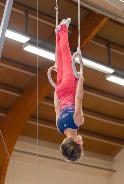 Young male gymnast performs an inverted hang on still rings, body fully extended, in an indoor gymnastics hall.