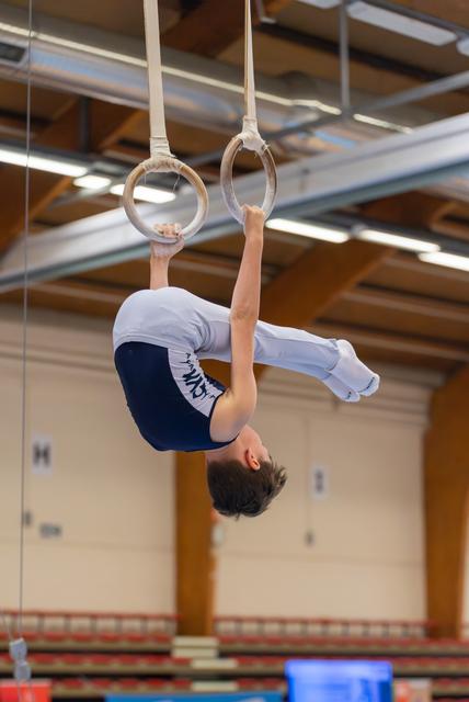 Young male gymnast performs an inverted hang on still rings, body curved with concentration, inside a sports hall.