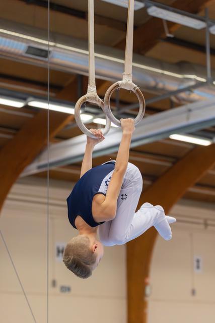 Young male gymnast hangs inverted on still rings, body tucked, showing intense focus and strength in a gymnasium.