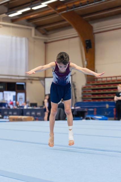 Young gymnast mid-air on floor mat, arms spread wide, focused gaze downward, ankle bandaged, indoor gymnasium.