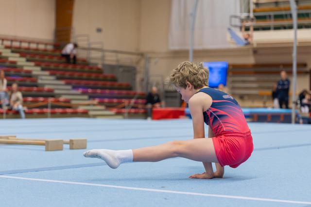Young boy gymnast performs a leg extension on the blue floor mat, leaning forward with concentration during a gymnastics meet.