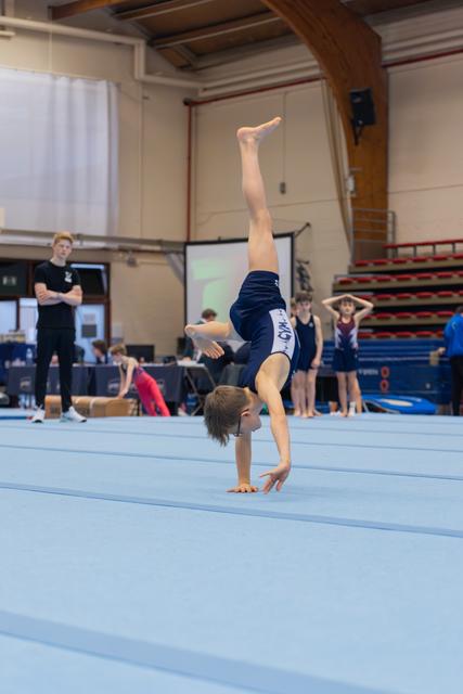 Young male gymnast balances on one hand during floor exercise, legs extended skyward, coaches watching in background.
