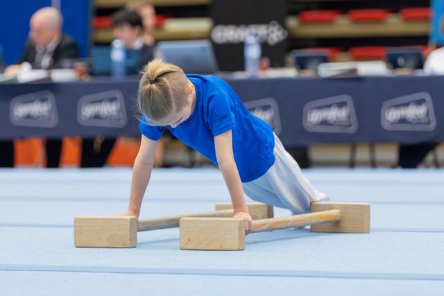 Young child in blue shirt performs parallel bar exercise on blue mat, leaning forward with intense concentration.