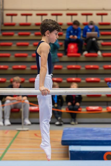 Young male gymnast grips parallel bars with focused concentration, wearing navy leotard and white trousers in a gymnasium.
